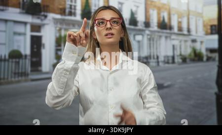 Junge blonde Frau zeigt mit dem Finger auf der Straße nach oben, während sie rote Brille und weißes Hemd trägt; Entschlossenheit. Stockfoto
