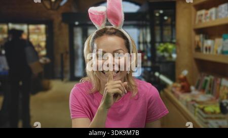 Frau in rosa Hemd mit Hasenohren lächelt und hält den Finger an die Lippen, um im Gebäude Ruhe zu finden; ruhige Geheimhaltung. Stockfoto