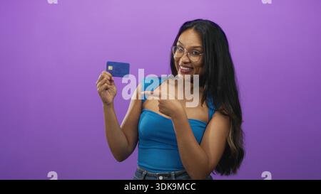 Frau, die eine Kreditkarte hält und mit dem Finger auf die Karte zeigt, im violetten Hintergrund des Studios, zeigt Chip und lächelnd; finanzielles Vertrauen. Stockfoto