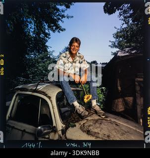 Ein glücklicher Mann sitzt beiläufig auf dem rostigen Dach eines alten VW-Käfers, der eine Sonnenblume hält. Stockfoto