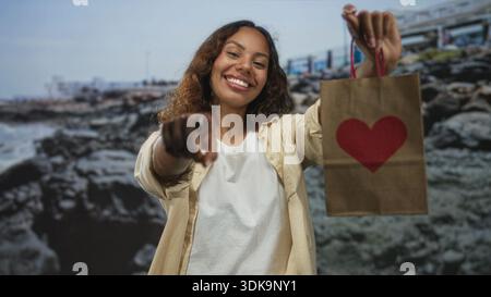Frau zeigt mit dem Finger auf die Kamera, während sie eine Papiertüte mit rotem Herzen an der Küste hält; Freude schenken. Stockfoto