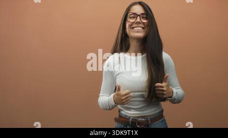 Frau mit langen Haaren und Brille zeigt zwei Daumen nach oben, zeigt sichtbare Hände und ein breites Lächeln, trägt ein weißes Hemd, Jeans und Gürtel in der Studiowand Stockfoto