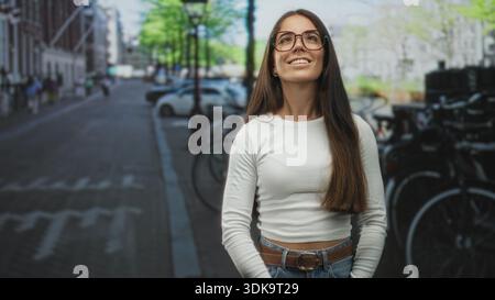 Frau mit Brille lächelt mit geschlossenen Augen und Händen locker an der Taille auf einer Straße neben geparkten Fahrrädern und Autos; ruhige Zufriedenheit. Stockfoto
