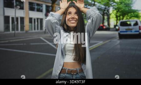 Frau hebt die Hände über den Kopf und zeigt Palmen und lächelt auf der Straße vor dem Gebäude, trägt weißes Crop Top und Jeans aus Denim; Freude verspielter Optimismus. Stockfoto