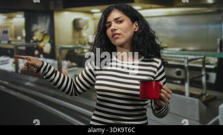 Junge brünette Frau zeigt mit dem Finger nach vorne, während sie im Küchengebäude einen roten Becher hält; Missbilligung. Stockfoto