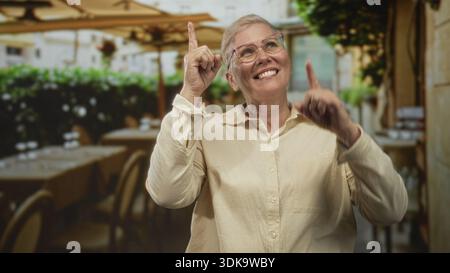 Die Frau zeigt mit den Zeigefingern auf der Straßenterrasse neben den Kaffeetischen, trägt eine Brille und lächelt; Freude feiern. Stockfoto