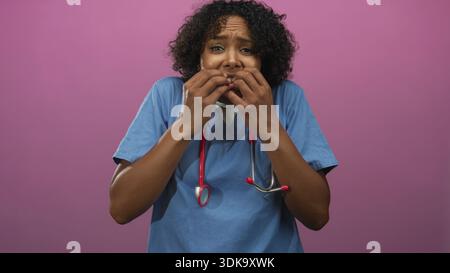 Junge afroamerikanische Ärztin mit blauen Peelings mit Stethoskop beißt Nägel im Studio; Angst. Stockfoto