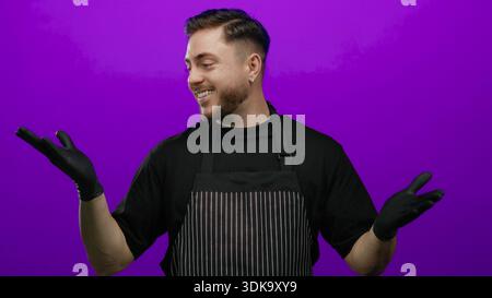 Junger Mann mit Bart, Schürze und Handschuhe, lächelnd vor isoliertem lila Hintergrund, mit einem fröhlichen Kellnerlook. Stockfoto