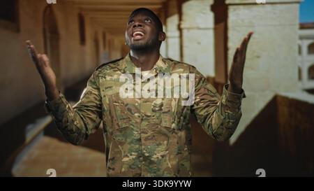 Mann Soldat in Tarnuniform mit überkreuzten Armen und sichtbaren Händen, grimmig in einem Steinbau-Korridor mit Bögen und Säulen; Dienstreflex Stockfoto