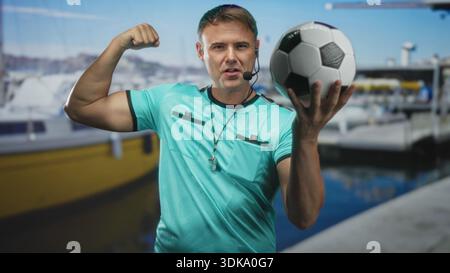 Der Schiedsrichter hält Fußball und Pfeife und bizepst im Studio, wobei das Headset-Mikrofon sichtbar ist; Stolz auf Selbstvertrauen. Stockfoto