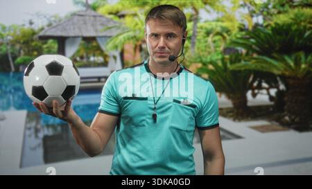 Mann Schiedsrichter hält Fußball in der rechten Hand mit Pfeife und Headset am Poolgebäude; Entscheidungsbefugnis. Stockfoto