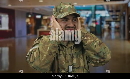 Hispanischer Mann in Tarnuniform bedeckt die Ohren mit beiden Händen inmitten des überfüllten Einkaufszentrums; Unwohlsein. Stockfoto