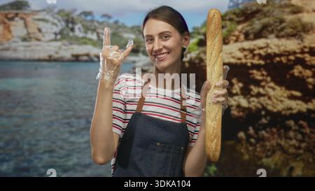 Frau mit Schürze und Kunststoffhandschuhen, die ein langes Baguette hält und im Studio Geste mit Steinhörnern zeigt; Freude. Stockfoto