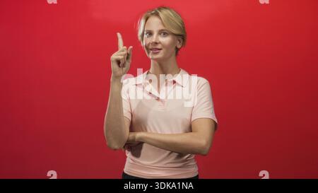 Frau zeigt den Finger nach oben mit gefaltetem Arm in der roten Wand des Studios, Zeigefinger als Geste hochgehoben; Selbstbewusstsein. Stockfoto