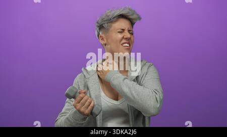 Frau mit kurzen Haaren in grauem Hoodie, die Hantel hält und den Hals im lila Studio umklammert; Unwohlsein. Stockfoto