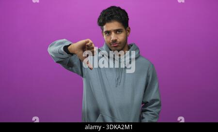 Mann in grauem Hoodie zeigt Daumen nach unten mit sichtbarem Daumen und neutralem Ausdruck im Studio; Ablehnung der Ablehnung. Stockfoto