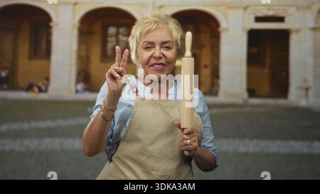 Die Köchin hält eine Rollnadel und zeigt mit der Hand ein Friedenszeichen im Innenhof des Gebäudes; Freude beim Backen. Stockfoto