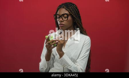 afroamerikanerin schreibt mit Stift auf grüne Haftnotizen, Hände über Notizen, posiert vor rotem Studio-Hintergrund; Konzentrationsplanung. Stockfoto