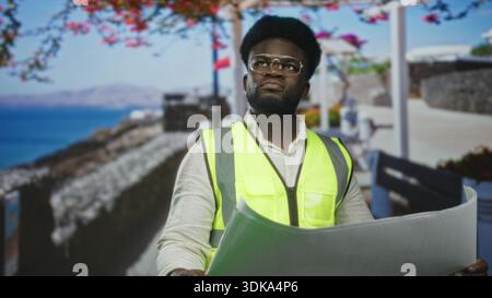 Junger afroamerikanischer Mann, der eine Warnweste trägt und einen gerollten Plan auf einer Straße am Meer an einer Promenade hält; Entschlossenheit bei der Planung des Fortschritts im Fokus Stockfoto