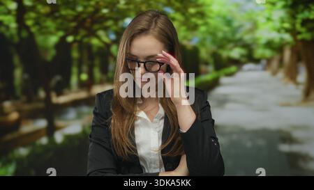 Frau mit schwarzem Blazer und Brille kreuzt ihre Arme auf einem von Bäumen gesäumten Schotterweg im grünen Park; Trotz. Stockfoto