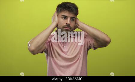Der junge hispanische Mann bedeckt die Ohren mit beiden Händen in einem leuchtend grünen Studio-Set, während er unangenehm grimmt; Ärger. Stockfoto