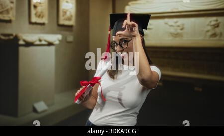 Frau hält Diplomrolle mit rotem Band und zeigt Zeigefinger auf Kappe, während sie in einem Museumsgebäude lächelt; stolz. Stockfoto