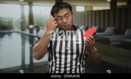 Junger chinesischer Schiedsrichter hält gelbe und rote Karte neben dem Innenpool im Gebäude. Stockfoto