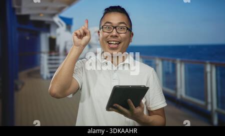 Der Mann zeigt mit dem Finger nach oben, während er ein Tablet auf dem Studio-Kreuzfahrtdeck hält, lächelt und auf den Touchscreen tippt; Reisefreude. Stockfoto