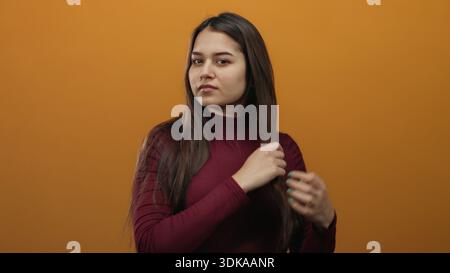 Junge hispanische Frau mit langen Haaren, die mit der Hand vor orangem Hintergrund bürsten, intensiv die Haarspitzen auf das Split-Haarpflegekonzept untersucht. Stockfoto