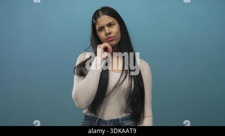 Lateinische Frau posiert mit Hand am Kinn im Studio vor blauer Wand, beugt sich leicht nach vorne und neigt den Kopf; Zweifel. Stockfoto