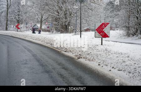 Rote und weiße Chevron-Warnschilder auf einer rutschigen kurvenreichen Straße, die mit Schnee bedeckt ist. Gefährliche Fahrbedingungen während eines schweren Schneesturms. Stockfoto