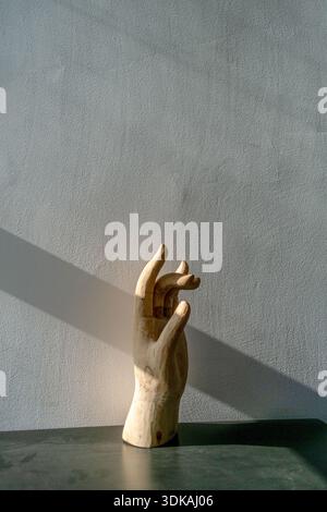 Künstlerische Handpuppen-Skulptur aus Holz in anmutiger Geste, die vor einer grauen Wand mit einem Sonnenstrahl steht und dramatische Schatten erzeugt. Stockfoto