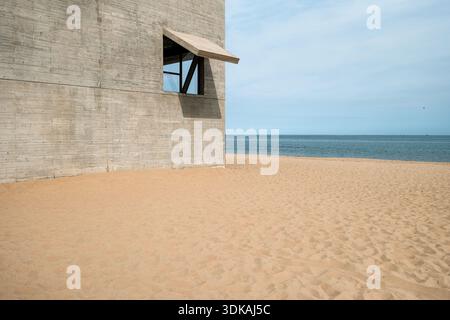 Ein minimalistisches Betonblockgebäude mit einem einzigen Fenster mit offenem Rolltor, das direkt an einem Sandstrand am Meer steht. Stockfoto