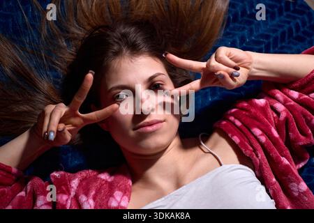 Der Schuss fängt eine junge Frau ein, die auf einer blauen Decke liegt, mit ihren Haaren um sie herum, sie macht spielerisch ein Friedenszeichen mit ihren Fingern und sie Stockfoto