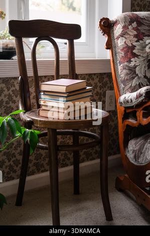 Stapel alter Bücher auf einem Holzstuhl vor dem Fenster mit Pflanzen Stockfoto