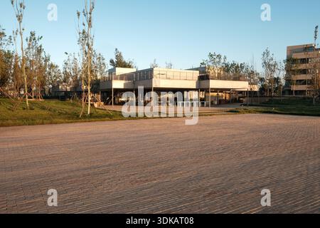 Weitwinkelblick auf ein modernes Gemeinschaftsgebäude aus Glas und Beton, umgeben von jungen Bäumen und einem von sanftem Abendlicht beleuchteten Schotterhof. Stockfoto