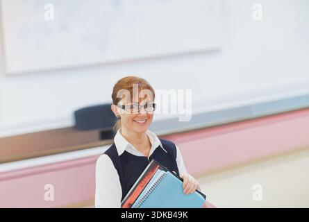 Reifer Lehrer, der in der Klasse an Bord steht, blaues Notizbuch hält, rote Mappe mit Bluse, Brille. Lehrer, Klassenzimmer, Podium, Journal, Ordner, sp Stockfoto