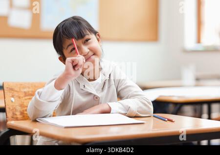 Asiatisches Mädchen mit hellgrauem Kapuzenpullover, das am Holzschreibtisch sitzt und ein Notizbuch mit rotem Bleistift hält. Klassenzimmer, Schüler, Lernen, Tageslicht, Konzentrat Stockfoto