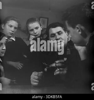 Slawjansk, Ukrainische SSR, Sowjetunion, 1960er Jahre Ein Archivfoto, das eine Nahaufnahme einer Berufsberatungsstunde zeigt. Ein dekorierter Offizier der sowjetischen Luftwaffe, ein Held der Sowjetunion, hält ein Modellflugzeug, während er mit einer Gruppe aufmerksamer Schulkinder spricht. Die Schüler, Jungen und Mädchen, hören mit großem Interesse den Geschichten des Veteranen zu. Die Goldstern-Medaille ist auf seiner Uniform sichtbar. Dieses Bild fängt die Atmosphäre der Mentorschaft, des Patriotismus und des Prestige des Militärberufs in der friedlichen Donbass-Region ein. Stockfoto