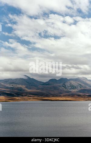 Ruhiger See, der weit entfernte Bergkämme unter einem breiten bewölkten Himmel reflektiert und ein friedliches und atmosphärisches Hochlandpanorama schafft. Stockfoto