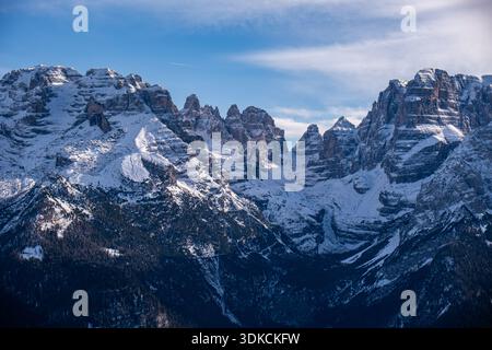Dramatische schneebedeckte Berggipfel der Dolomiten mit geschichteten Felsformationen, die sich über einem dunklen Alpenwald unter einem klaren blauen Winterhimmel erheben, Italien. Stockfoto