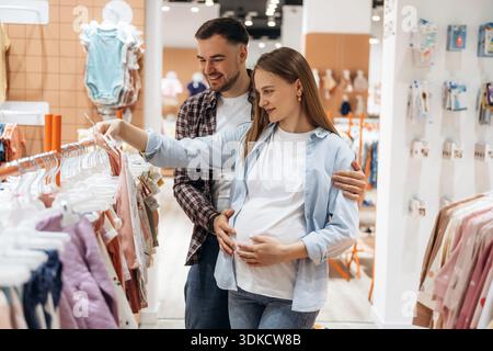 Ich fühle mich gut, wähle die Kleidung. Schwangere Frau mit Mann sind im Kinderbekleidungsgeschäft. Stockfoto