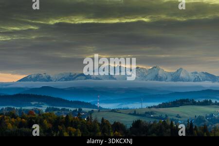 Panoramablick auf die schneebedeckten Tatra, die sich über sanften Hügeln und Wäldern unter dramatischem bewölktem Himmel in Südpolen erhebt. Stockfoto