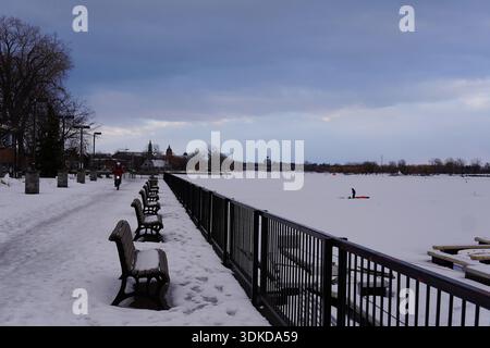 Perspektive auf leere Parkbänke, die im Winter an einem gefrorenen Flussufer mit Schnee bedeckt sind. Kalte Stadtlandschaft. Stockfoto
