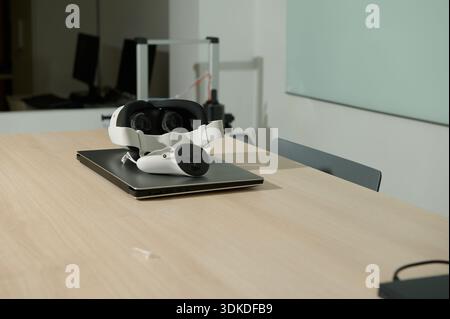Ein weißes VR-Headset und -Controller sitzen auf einem geschlossenen Laptop auf einem hellen Holztisch in einem ruhigen, modernen Arbeitsbereich, bereit für Vorführungen oder Schulungen. Stockfoto