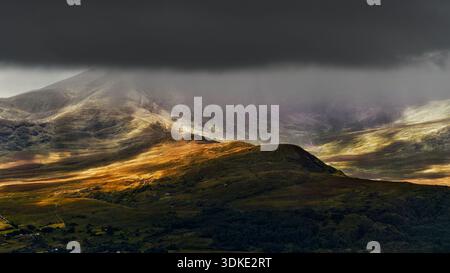 Die Sonne scheint durch die Wolken auf die grünen und braunen Hügel des Killarney National Park in Irland. Nebel und niedrige Wolken bedecken den fernen Moun Stockfoto