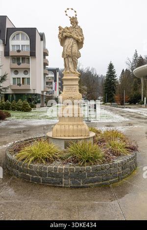 ROGASKA SLATINA, SLOWENIEN, 01.03.2026: Eine detailgetreue Steinstatue steht auf einem geschnitzten Sockel in einem kreisrunden Pflanzgefäß, umgeben von Grün in einer ruhigen Kuh Stockfoto