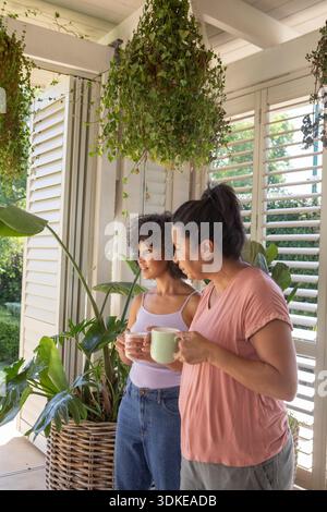 Mutter und Tochter stehen auf der Veranda neben den Fensterläden, halten Keramikbecher und bewundern den Hof, Kopierraum. Veranda, Grün, Entspannung, Verbundenheit, Inneneinrichtung, botanisches, gemütliches || Model veröffentlicht Stockfoto