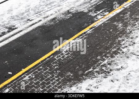 Ein schneebedeckter Stadtrand zeigt eine hellgelbe Linie, die dunklen Asphalt von einem gemauerten Bürgersteig trennt. Schnee bedeckt die Oberfläche und erzeugt eine kalte, strukturierte Oberfläche Stockfoto