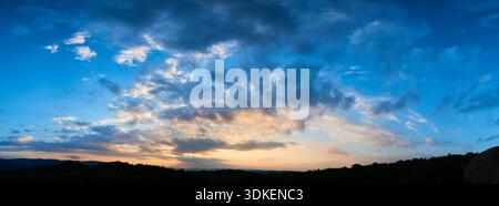 Atemberaubender Himmel voller dramatischer Wolken übergeht bei Sonnenuntergang von tiefem Blau zu sanften Pastelltönen. Horizont gesäumt von silbrigen Hügeln und Wäldern, die der ruhigen und weitläufigen Landschaft Tiefe verleihen. Stockfoto